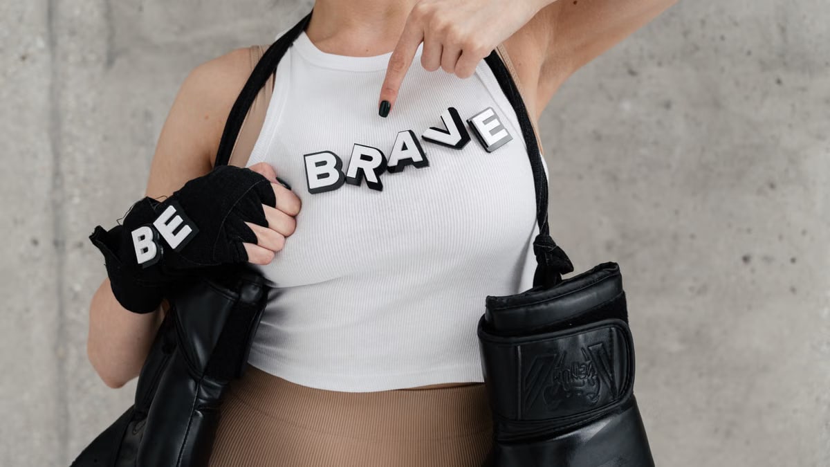 A woman points at 'Be Brave' letters on her shirt while wearing boxing gloves, conveying empowerment.